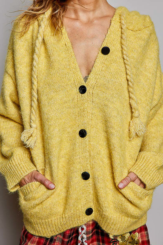 A close-up of a person wearing a chunky knit yellow cardigan with rope drawstrings, black buttons, and front pockets, paired with red plaid bottoms.