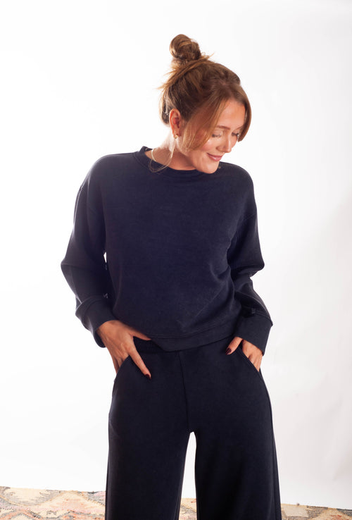 A woman wearing a dark navy blue crewneck sweatshirt and matching wide-leg sweatpants, standing against a plain white studio background.
