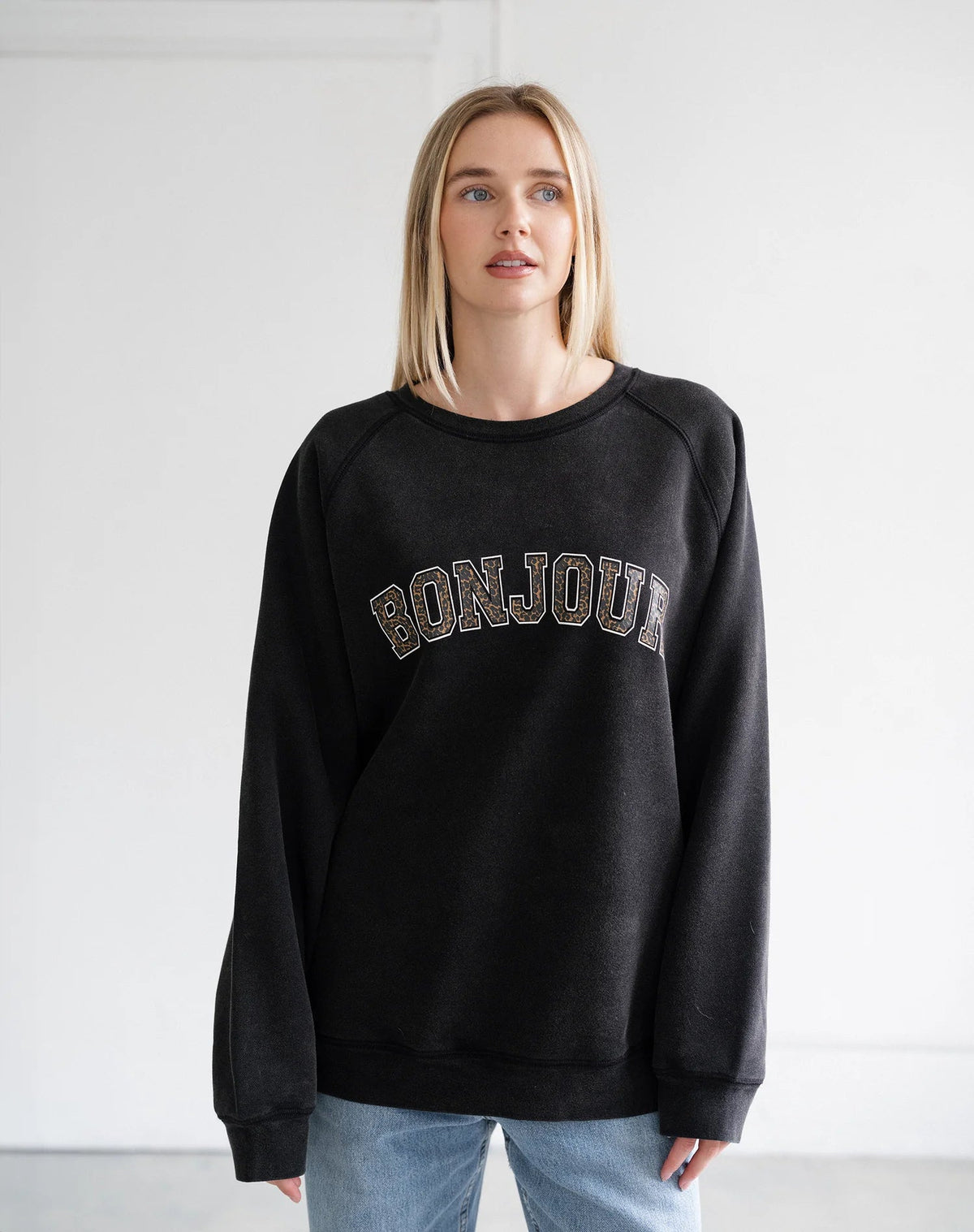 A blonde woman wearing a black crewneck sweatshirt with 'BONJOUR' written across the chest in a textured, metallic gold and silver pattern, paired with light blue jeans, standing against a plain white wall.