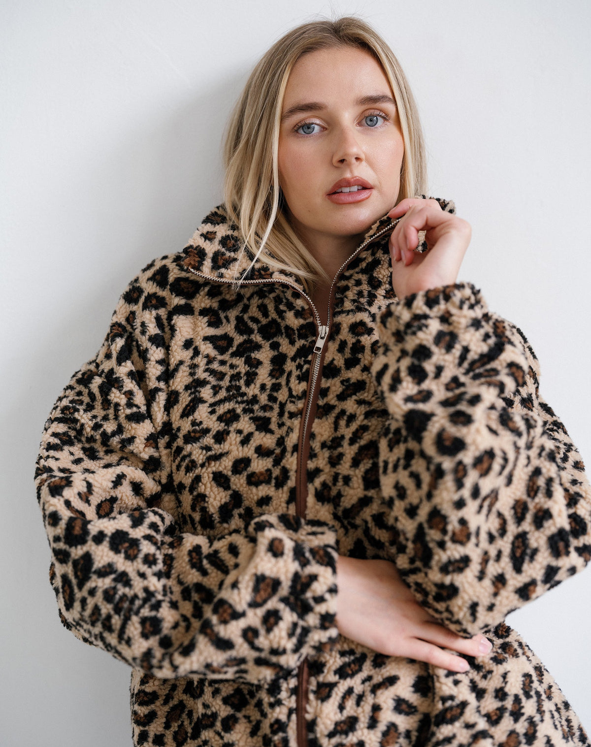 A woman wearing a plush leopard-print faux fur jacket with a high collar and front zipper, posed against a plain light gray wall.