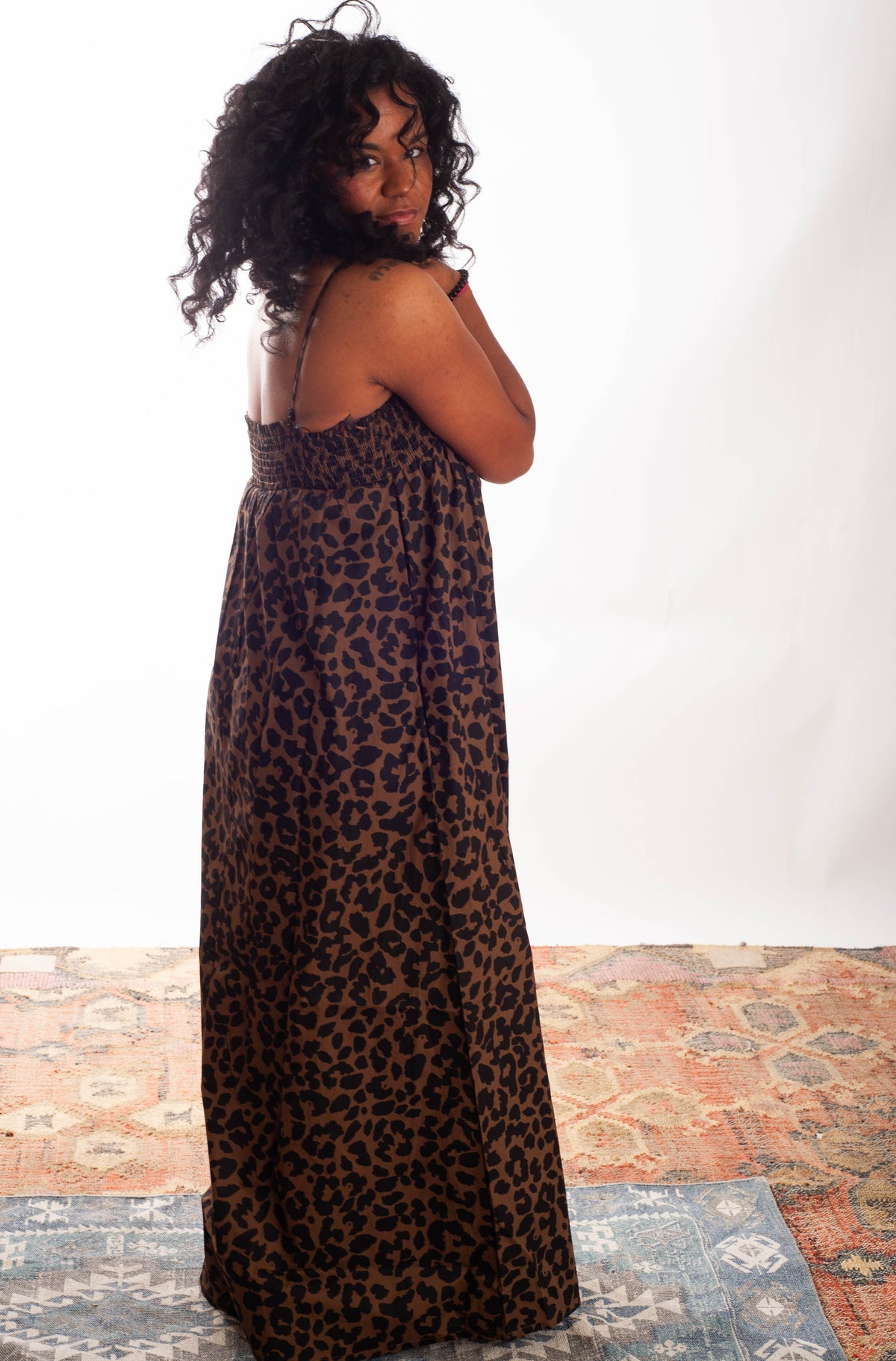 A woman wearing a leopard-print maxi dress, photographed from behind against a white studio background with a patterned rug on the floor.