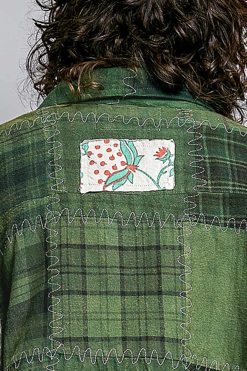 Close-up back view of a person wearing a green plaid flannel shirt with decorative zigzag stitching and a square floral patch on the upper back.