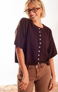 A woman wearing a dark brown short-sleeve button-up top and brown pants, smiling and looking down. She is wearing glasses and standing against a plain white background.