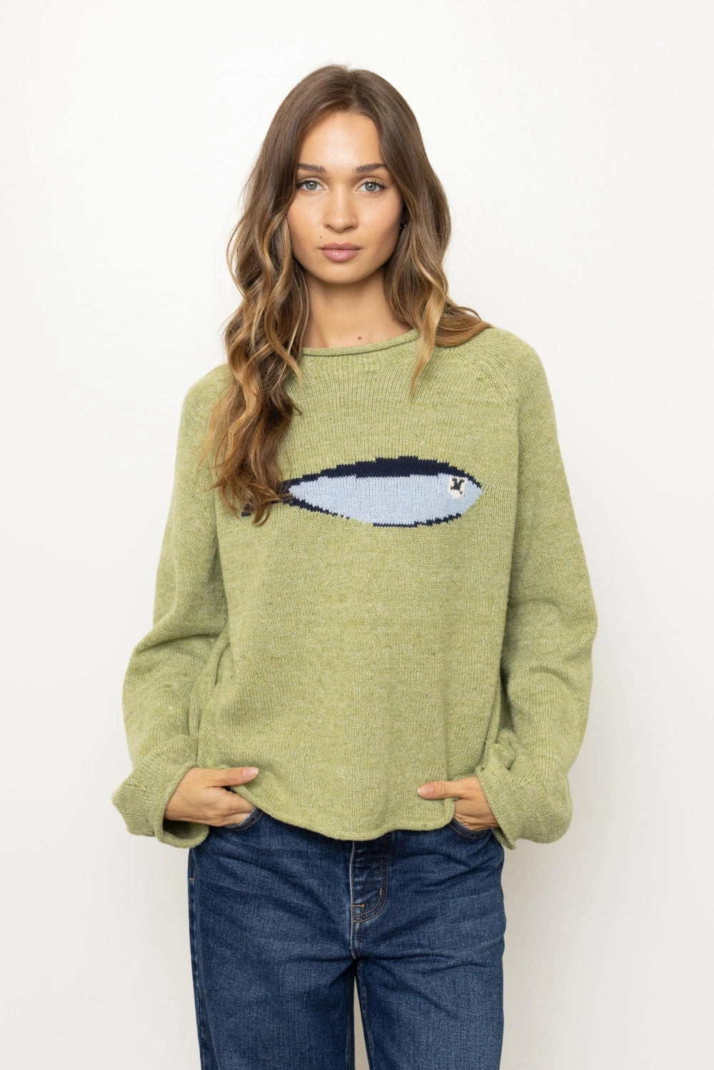 A woman modeling a light olive green knit sweater with a pixelated fish graphic across the chest, paired with dark blue jeans, against a plain white background.